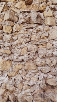 A textured stone wall composed of irregularly shaped rocks of varying sizes, closely packed together. The stones have a natural tan and grey color, displaying a rough, rustic appearance. Some gaps between the stones are filled with mortar, giving the structure an aged, historical feel.