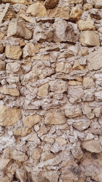 A textured stone wall composed of irregularly shaped rocks of varying sizes, closely packed together. The stones have a natural tan and grey color, displaying a rough, rustic appearance. Some gaps between the stones are filled with mortar, giving the structure an aged, historical feel.