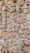 A textured stone wall composed of irregularly shaped rocks of varying sizes, closely packed together. The stones have a natural tan and grey color, displaying a rough, rustic appearance. Some gaps between the stones are filled with mortar, giving the structure an aged, historical feel.