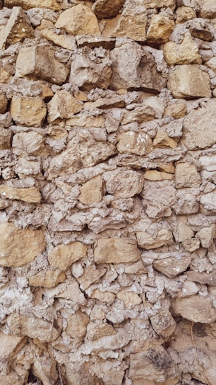 A textured stone wall composed of irregularly shaped rocks of varying sizes, closely packed together. The stones have a natural tan and grey color, displaying a rough, rustic appearance. Some gaps between the stones are filled with mortar, giving the structure an aged, historical feel.