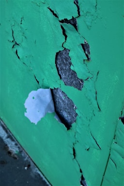 Peeling green paint reveals a rough, gray surface beneath. The paint is cracked and flaking, creating an abstract, textured pattern.