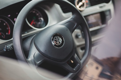 A close-up view of a car's interior focuses on the steering wheel, which has the Volkswagen logo in the center. The dashboard includes various gauges and controls, and the car's interior appears modern and clean.