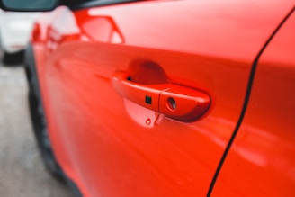 A close-up view of a red car door handle, highlighting its sleek design and glossy finish. The focus is on the handle while the backdrop is softly blurred, emphasizing the vibrant red color.