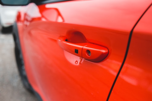 A close-up view of a red car door handle, highlighting its sleek design and glossy finish. The focus is on the handle while the backdrop is softly blurred, emphasizing the vibrant red color.