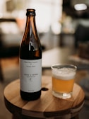 A bottle of Maine Beer Company Woods & Waters India Pale Ale is standing on a wooden surface next to a glass filled with the same beer. The label on the bottle is white with black text. The background is blurred, suggesting an indoor setting with soft lighting.