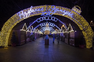 A festive outdoor market scene at night, featuring archways adorned with glowing yellow and blue Christmas lights. The path is lined with wooden stalls decorated with additional lights, while people walk through the illuminated area, creating a warm and inviting atmosphere.