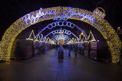 A festive outdoor market scene at night, featuring archways adorned with glowing yellow and blue Christmas lights. The path is lined with wooden stalls decorated with additional lights, while people walk through the illuminated area, creating a warm and inviting atmosphere.