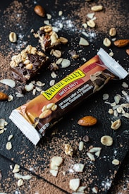 A Nature Valley Sweet & Salty snack bar is displayed on a dark textured surface surrounded by scattered almonds, peanuts, oats, and cocoa powder. The bar is wrapped in packaging that features images of chocolate and almonds.