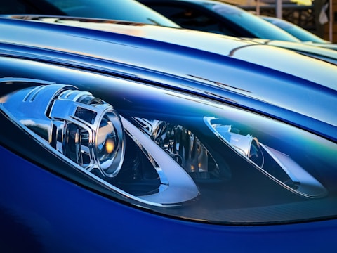 Close-up of a sleek and modern car headlight featuring intricate design elements and reflective surfaces. The surrounding car body is smooth and glossy, with a metallic blue finish that enhances its stylish appearance. The lighting offers a dramatic and detailed view, highlighting the craftsmanship of the headlight components.
