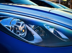 Close-up of a sleek and modern car headlight featuring intricate design elements and reflective surfaces. The surrounding car body is smooth and glossy, with a metallic blue finish that enhances its stylish appearance. The lighting offers a dramatic and detailed view, highlighting the craftsmanship of the headlight components.