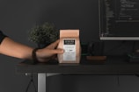 A hand is holding a small brown paper bag labeled 'Oaxaca café orgánico' over a dark desk. Next to it sits a dark green mug on a coaster. In the background, a computer monitor displays lines of code. A small potted plant is also visible, adding a touch of green to the setting.