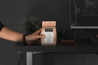 A hand is holding a small brown paper bag labeled 'Oaxaca café orgánico' over a dark desk. Next to it sits a dark green mug on a coaster. In the background, a computer monitor displays lines of code. A small potted plant is also visible, adding a touch of green to the setting.
