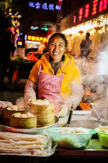 A smiling vendor wearing a yellow jacket and a red checkered apron is standing at a food stall. The stall has steam rising from bamboo steamers filled with dumplings. There are containers of various ingredients on the counter. In the background, bright neon signs and lights create a festive atmosphere.