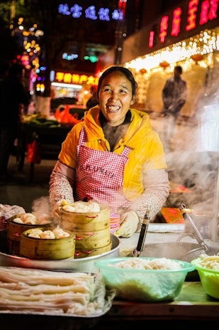 A smiling vendor wearing a yellow jacket and a red checkered apron is standing at a food stall. The stall has steam rising from bamboo steamers filled with dumplings. There are containers of various ingredients on the counter. In the background, bright neon signs and lights create a festive atmosphere.