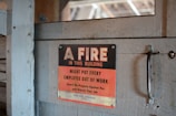 A worn, metal sign mounted on a wooden surface reads, 'A FIRE IN THIS BUILDING MIGHT PUT EVERY EMPLOYEE OUT OF WORK. Guard the Property Against Fire and Protect Your Job.' The sign features a bold black and red color scheme with a rustic backdrop.