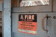 A worn, metal sign mounted on a wooden surface reads, 'A FIRE IN THIS BUILDING MIGHT PUT EVERY EMPLOYEE OUT OF WORK. Guard the Property Against Fire and Protect Your Job.' The sign features a bold black and red color scheme with a rustic backdrop.