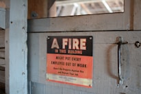 A worn, metal sign mounted on a wooden surface reads, 'A FIRE IN THIS BUILDING MIGHT PUT EVERY EMPLOYEE OUT OF WORK. Guard the Property Against Fire and Protect Your Job.' The sign features a bold black and red color scheme with a rustic backdrop.