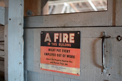 A worn, metal sign mounted on a wooden surface reads, 'A FIRE IN THIS BUILDING MIGHT PUT EVERY EMPLOYEE OUT OF WORK. Guard the Property Against Fire and Protect Your Job.' The sign features a bold black and red color scheme with a rustic backdrop.