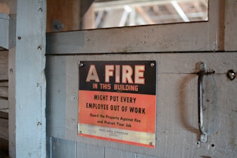 A worn, metal sign mounted on a wooden surface reads, 'A FIRE IN THIS BUILDING MIGHT PUT EVERY EMPLOYEE OUT OF WORK. Guard the Property Against Fire and Protect Your Job.' The sign features a bold black and red color scheme with a rustic backdrop.