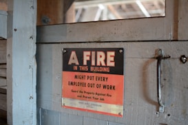 A worn, metal sign mounted on a wooden surface reads, 'A FIRE IN THIS BUILDING MIGHT PUT EVERY EMPLOYEE OUT OF WORK. Guard the Property Against Fire and Protect Your Job.' The sign features a bold black and red color scheme with a rustic backdrop.