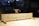 A wooden sign with handwritten words stating 'The Carpenter is out for tea!!' is situated in a workshop with blurred background elements suggesting tools and woodworking equipment.