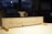 A wooden sign with handwritten words stating 'The Carpenter is out for tea!!' is situated in a workshop with blurred background elements suggesting tools and woodworking equipment.