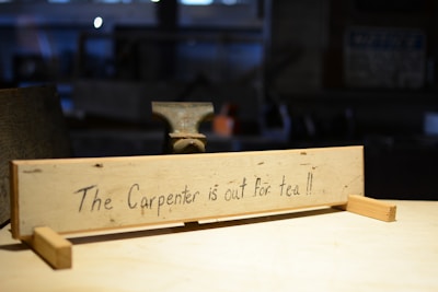 A wooden sign with handwritten words stating 'The Carpenter is out for tea!!' is situated in a workshop with blurred background elements suggesting tools and woodworking equipment.