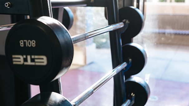 A set of gym weights is stacked on a rack near a bright window. The weights are arranged in descending order, with shiny metal bars connecting them. Natural light filters through the window, creating a calm and focused atmosphere.