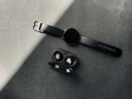 A modern black smartwatch with a sleek design is displayed on a textured surface next to a pair of wireless earbuds in an open case. The lighting creates subtle contrasts and highlights the smooth matte finish of the items.