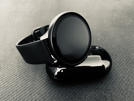 A sleek, modern smartwatch with a round face displayed against a textured, dark surface. The watch is placed atop a glossy, oval-shaped object, possibly a case or charger.