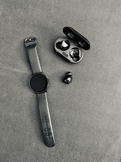 A black smartwatch with a rubber strap is placed on a textured fabric surface next to a pair of wireless earbuds. The earbuds are accompanied by their open charging case, which is also black.