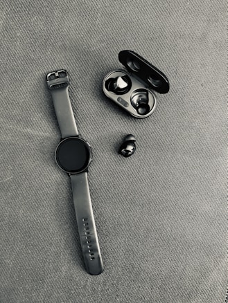 A black smartwatch with a rubber strap is placed on a textured fabric surface next to a pair of wireless earbuds. The earbuds are accompanied by their open charging case, which is also black.