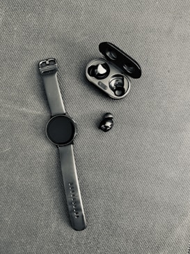 A black smartwatch with a rubber strap is placed on a textured fabric surface next to a pair of wireless earbuds. The earbuds are accompanied by their open charging case, which is also black.