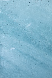 A textured surface with varying shades of blue, resembling weathered or painted concrete. Small black speckles are scattered across the surface, adding to the rough appearance. The color transitions subtly between lighter and darker hues, giving it a somewhat marbled effect.