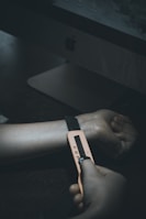 A dark, moody image depicting a person with a strap or device wrapped around their wrist, seemingly placed there by another person's hands. The focus is on the hand and wrist area, with a computer monitor visible in the background.