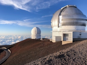Two large astronomical observatories with dome-shaped structures are situated on a barren, rocky mountain top under a clear blue sky filled with scattered clouds. A winding road leads up to the observatories.