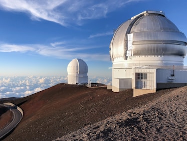 Two large astronomical observatories with dome-shaped structures are situated on a barren, rocky mountain top under a clear blue sky filled with scattered clouds. A winding road leads up to the observatories.
