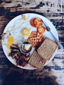 A hearty breakfast plate contains two fried eggs with slightly runny yolks, sliced roasted tomatoes, baked beans, sautéed mushrooms, crispy bacon, and slices of toasted bread. A small dish of butter is included, and the meal is served on a rustic wooden tabletop with a fork and knife placed beside the plate.