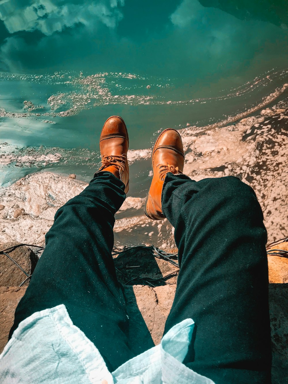 person in black pants and brown leather shoes sitting on rock near body of water during