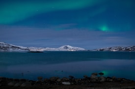 A serene landscape featuring a calm body of water reflecting the lights of a small settlement along its far shore. Snow-capped mountains form a majestic backdrop, while the sky is adorned with soft, ethereal streaks of the northern lights.