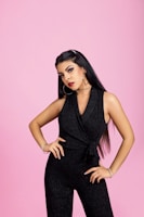 A woman stands confidently against a pink background wearing a black sleeveless jumpsuit. She has long dark hair and is wearing large hoop earrings, a necklace, and bracelets. Her makeup is bold, with striking eye makeup and red lipstick, and she poses with one hand on her hip.