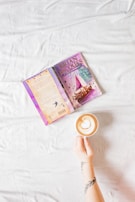An open book with a colorful cover lies on a white fabric background next to a hand holding a cup of coffee. The book's cover features an illustration of a ship, and the hand is tattooed. The cappuccino has an intricate heart-shaped latte art on the surface.