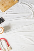 A white fabric background features a wooden cutting board, a cracked smartphone, two rings, a ceramic cup with a latte, and a woven rainbow decoration in red and white.