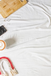 A white fabric background features a wooden cutting board, a cracked smartphone, two rings, a ceramic cup with a latte, and a woven rainbow decoration in red and white.