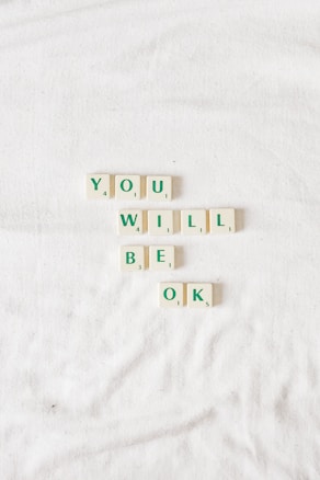 Scrabble tiles on a soft, white surface form a reassuring message. The words are arranged in two rows, creating an uplifting phrase using simple typography.