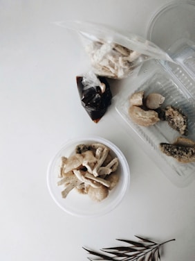 Various food items are arranged on a white surface, including a clear plastic bowl containing light-colored pieces of fried snacks, a transparent plastic container with pieces of food that appear to be seasoned, and a small plastic bag filled with a dark liquid. A twig with leaves is placed in the lower corner.