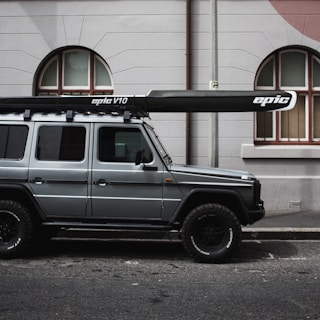 A gray off-road vehicle is parked on a street next to a building with arched windows. A long black and white kayak labeled 'epic V10' is mounted on the roof of the vehicle.