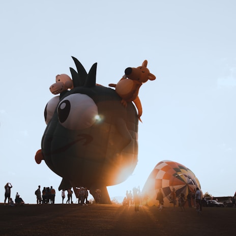 A large, whimsical hot air balloon shaped like a cartoonish green character with big eyes, and additional characters resembling a kangaroo and a hippo on top. The scene is set during a sunset, casting a warm glow and long shadows on a grassy field where several people are gathered, some taking photos.