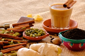 A glass of chai sits on a saucer with cinnamon sticks inside, surrounded by various spices including cardamom pods, ginger, and ground tea leaves. The spices are arranged on a burlap surface, creating a rustic and aromatic ambiance.