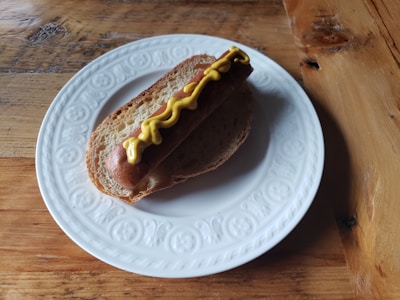 A hot dog is placed in a sliced piece of bread on a white decorative plate. The hot dog is topped with a wavy line of yellow mustard. The plate rests on a wooden table, which has a rustic appearance with visible grain and texture.